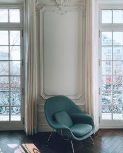 Photo d'un siège vert devant un magnifique mur avec ses moulures et entre 2 grandes fenêtres donnant sur l'extérieur parisien